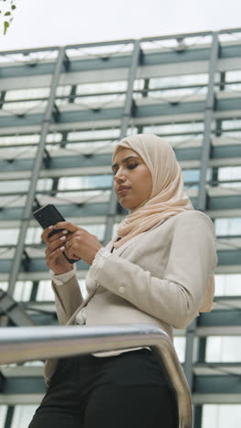 Vertikales-Video-Einer-Muslimischen-Geschäftsfrau,-Die-Nachrichten-Auf-Dem-Mobiltelefon-überprüft,-Während-Sie-Vor-Dem-Büro-In-Der-Stadt-Steht